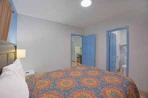 A bedroom with a patterned bedspread, a bedside lamp, and a door leading to a bathroom. - Queen Bed|Kitchenette|Non-Smoking (Merritt Island FL)