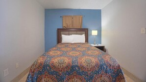 A bedroom with a patterned bedspread, a wooden headboard, a nightstand with a lamp, and a window with curtains. - Queen Bed|Kitchenette|Non-Smoking (Merritt Island FL)