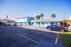 A parking lot with several cars, a blue building with a flat roof, and palm trees in the background. - 2 Full Beds|Non-Smoking (Merritt Island FL)