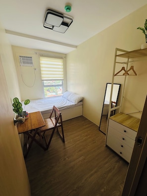 A compact room with a bed, a desk, a chair, a hanging clothes rack, and a window with blinds.