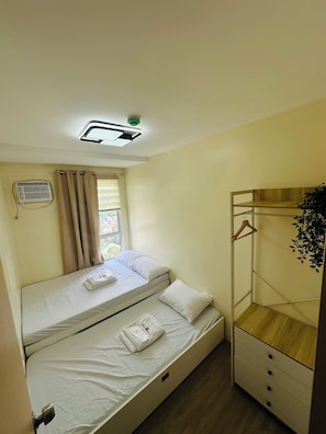 Un lit simple avec un matelas et des oreillers blancs, une table de chevet agrémentée d’une plante suspendue, un climatiseur fixé au mur et une fenêtre avec des rideaux.
