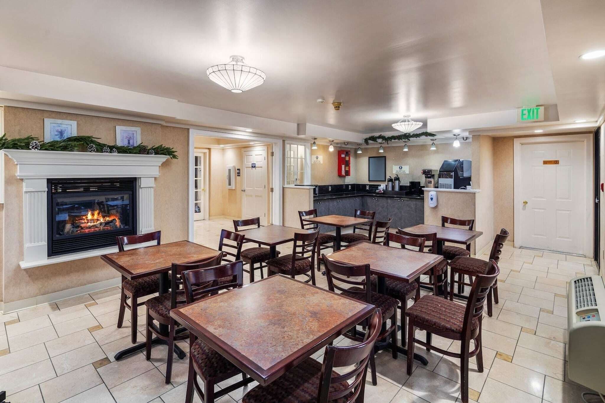 A hotel lobby with a fireplace, multiple tables and chairs, a kitchen area, and an exit sign.