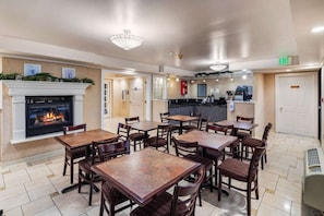 A hotel lobby with a fireplace, multiple tables and chairs, a kitchen area, and an exit sign.