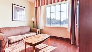 Ein Zimmer mit einer Couch, einem Tisch, einem fensterbelaubten Fenster, einer Lampe und einem gerahmten Bild an der Wand.
