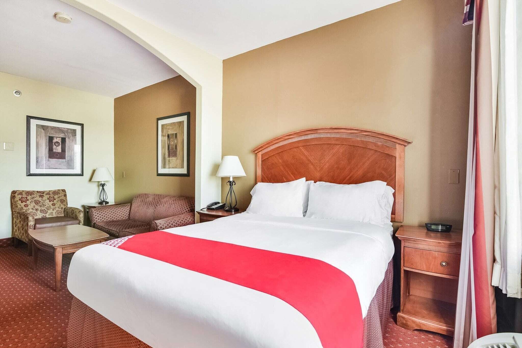 A hotel room with a large bed, a wooden headboard, a desk, a chair, two lamps, and framed pictures on the walls.