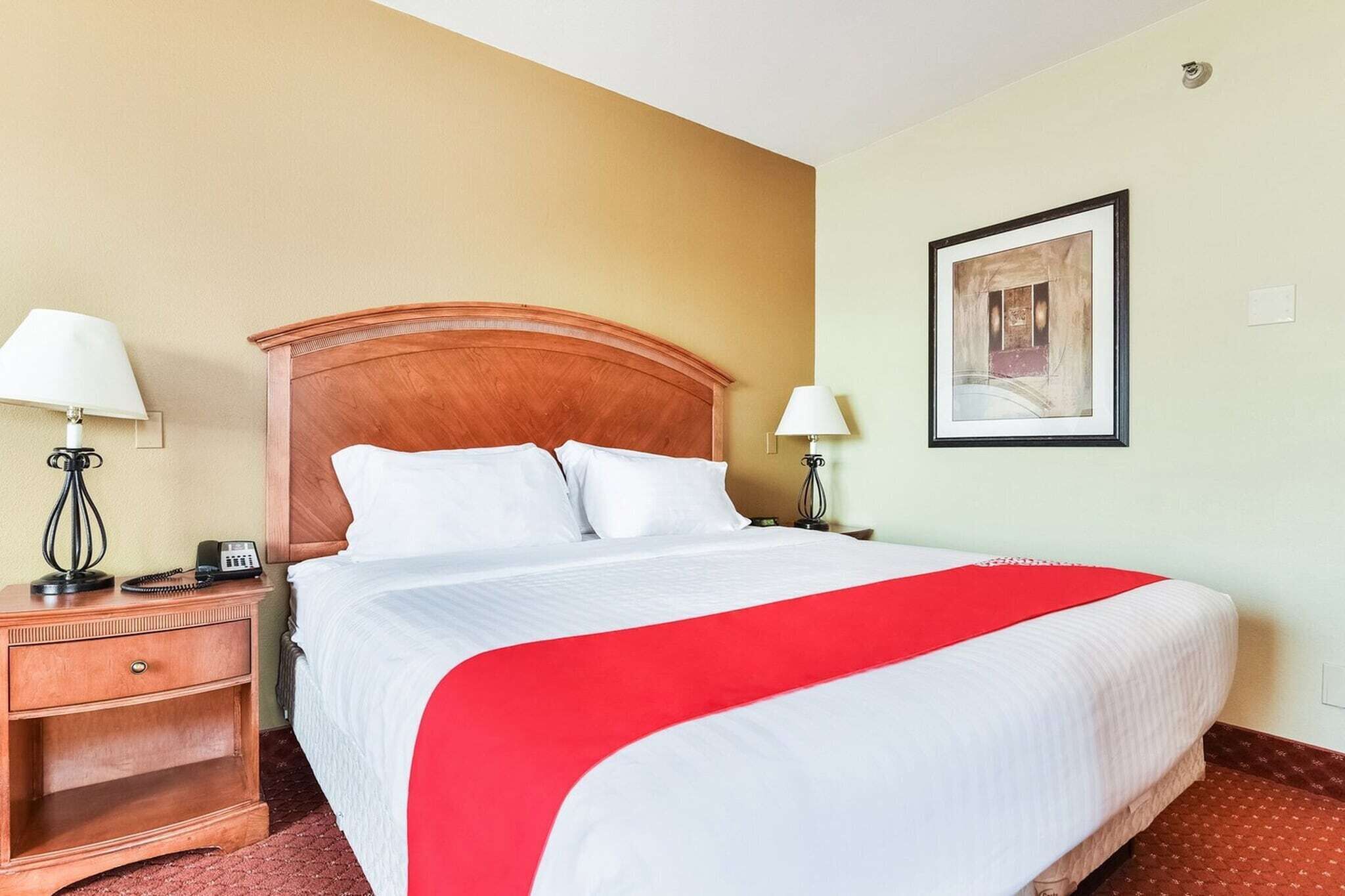 A hotel room with a wooden headboard, a large bed with white bedding and a red accent, two bedside tables with lamps, and a framed picture on the wall.