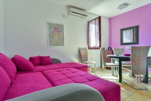 Una habitación con un sofá rosa, una mesa de comedor con sillas, un ventanal con cortinas y un equipo de aire acondicionado en la pared.