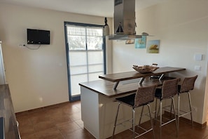 Eine Küche mit Fernseher, einem Fenster mit Jalousien, einer Bar mit Hockern und einer kleinen Kochinsel.