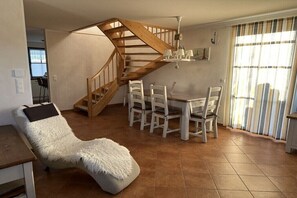 Ein Zimmer mit einer Holztreppe, einem Esstisch mit Stühlen, einem gemütlichen Sessel und einem Fenster mit Vorhängen.
