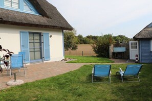 Ein Gebäude mit einem Reetdach, heller Fassade und blauen Fensterläden, eine gepflasterte Terrasse mit Sitzgelegenheiten und eine Rasenfläche.