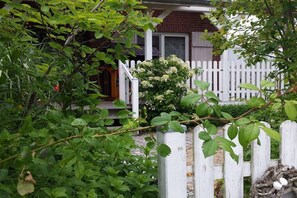 Ein weißer Holzzaun, ein Backsteinhaus und ein Garten mit grünem Laub und weißen Blüten.