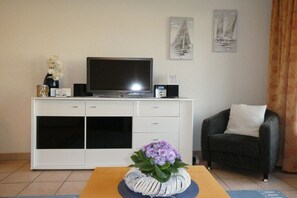 Ein Wohnzimmer mit einem Flachbildfernseher auf einem weißen Schrank, einem dunklen Sessel und einem Tisch mit einer Blumenvase.