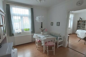 Ein Zimmer mit Esstisch, Stühlen, einem Bücherregal, einer Uhr und einem Fenster mit Vorhängen.