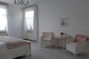 Ein Schlafzimmer mit einem Bett, zwei Stühlen, einem kleinen Tisch, einer Chandelier und einem Fenster mit Vorhängen.