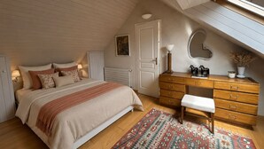 Ein Schlafzimmer mit schräg versiegelter Decke, einem Bett mit gemustertem Bettbezug, einer Holzkommode, einer Bank, einem Teppich mit Muster und einem an der Wand befestigten Spiegel.