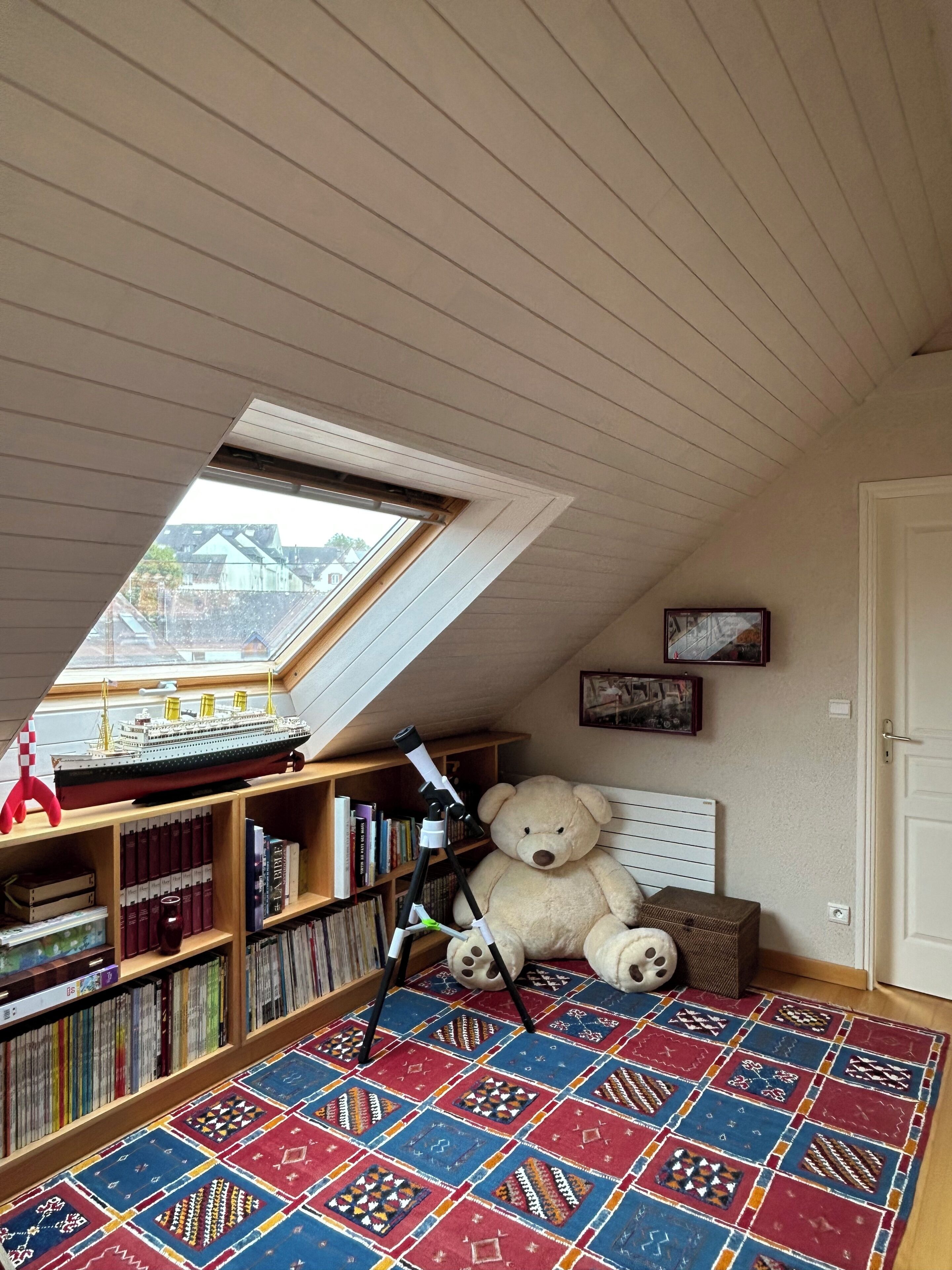 A room with a sloped ceiling, a skylight, a large teddy bear, a bookshelf, and a patterned rug.