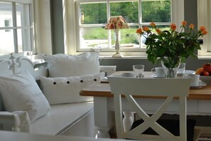 Ein Zimmer mit einem weißen Sofa, einem Tisch mit Lampe, einer Vase mit Blumen und Blick auf Grünfläche draußen.