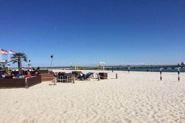 Ein Strand mit Volleyballnetz, Liegestühlen und einem Steg im Hintergrund.