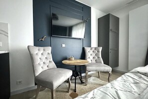 Ein modernes Hotelzimmer mit einem Bett, zwei Polstersesseln, einem kleinen runden Tisch, einem Flachbildfernseher an einer dunklen Wand und einem Kleiderschrank.