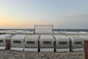 Ein Strand mit Reihen weißer Stühle, die mit 'KINO' beschriftet sind, und einer großen Leinwand im Hintergrund.