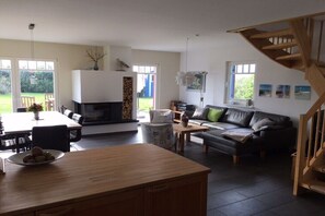A modern living room with a fireplace, a dining area, a staircase, and large windows. - Holiday home with 2 bedrooms (Putgarten)