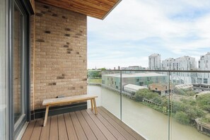 Uma varanda com um banco de madeira, um muro de tijolos e vista para um rio e edifícios.