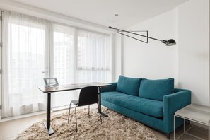 Ein modernes Wohnzimmer mit einem salbeingrünfarbenen Sofa, einem Glastisch, einem schwarzen Sessel, einem weißen Teppich und einer Stehlampe.