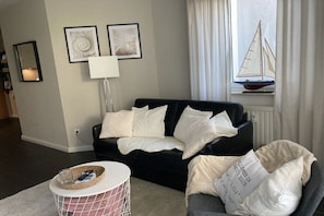 A living room with a black sofa, a round coffee table, a lamp, and a window with a sailboat decoration. - Holiday apartment with 1 bedroom (Kühlungsborn)