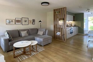 A modern living room with a grey sectional sofa, a small white coffee table, and a kitchen area with white cabinets and appliances. - Forest settlement "Time Out" (Prerow)