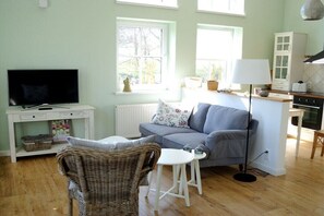 Ein Wohnzimmer mit einer Couch, einem Fernseher, einem Korbsessel, einem kleinen Tisch und einer Lampe.