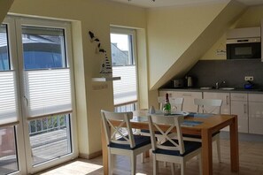Ein Essbereich mit Tisch und Stühlen, eine Küche mit Geräten sowie ein Fenster mit Jalousien.
