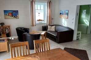Ein Wohnzimmer mit einer Couch, einem Esstisch mit Stühlen, einem Bücherregal und zwei Bildern an der Wand.