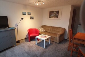 Ein Wohnzimmer mit einer Couch, einem roten Sessel, einem kleinen weißen Tisch, einem Fernseher, einer Stehlampe und einem Bild an der Wand.