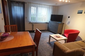 Ein Wohnzimmer mit einem Holztisch, einem roten Sessel, einer Couch, einem Fernseher und einem Fenster mit Jalousien.