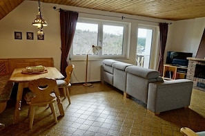 Ein gemütliches Zimmer mit Holzbalkendecke, einer Couch, einem Fernseher, einem Kamin und einer Essecke mit Tisch und Stühlen.