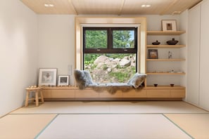 A room with tatami flooring, a window with a view of rocks and greenery, a wooden shelf with decorative items, and a small stool with figurines. - Koi by H2 Life (Niseko)