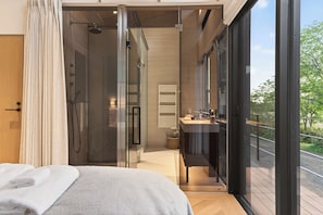 A modern bedroom with a large bed, a glass shower enclosure, a bathroom with a sink, and a balcony with a view.
