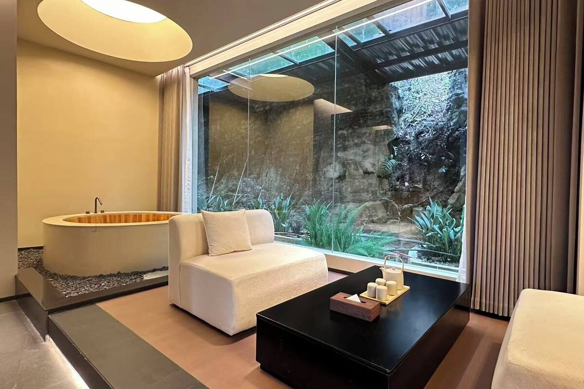 Ein modernes Badezimmer mit einer Badewanne, einem Sofa, einem Couchtisch und Blick auf einen felsigen Garten.