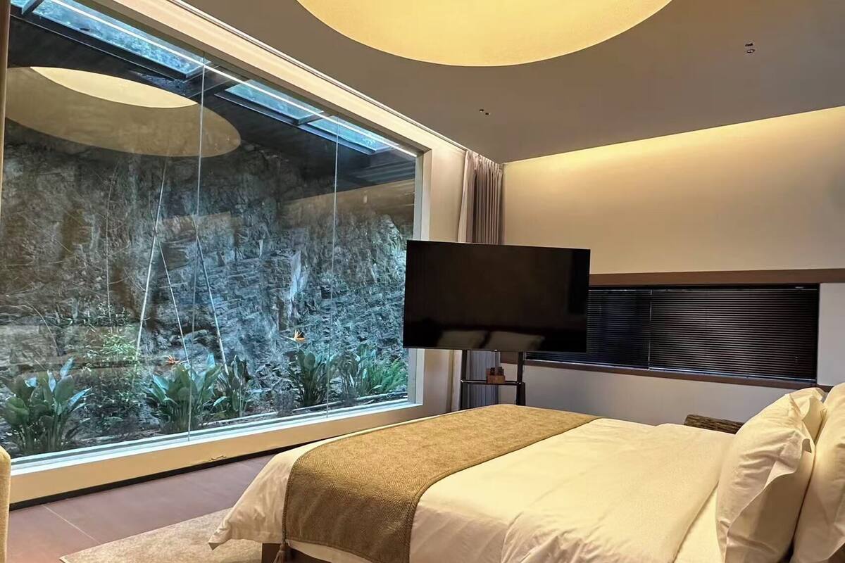 A bedroom with a large window, a bed, a flat-screen TV, and a stone wall view.