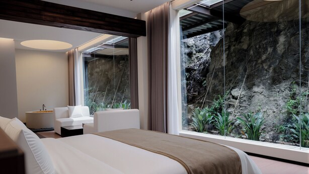 A modern hotel room with a large bed, a sitting area with a sofa and armchairs, a small table with a lamp, and a view of a rocky landscape through large windows.