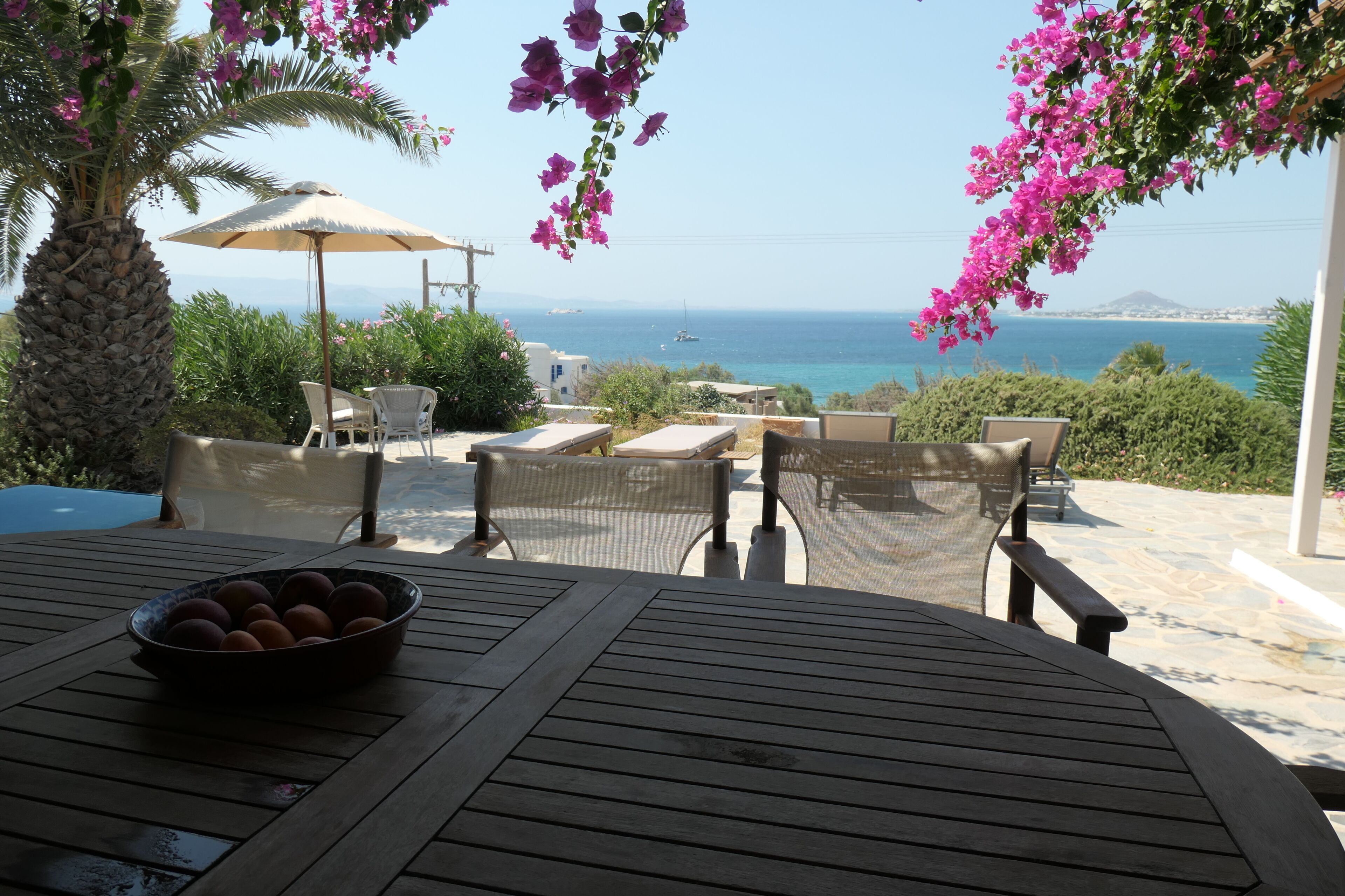 A wooden table with a bowl of fruit, outdoor seating, and a view of the sea.