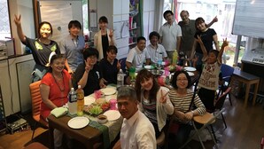 A group of people gathered around a table with food and drinks, in a room with a whiteboard, windows, and a door.
