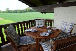 Ein Holztisch für vier Personen, ausgestattet mit karierten Kissen, mit Blick auf ein grünes Feld und einem Balkongeländer.