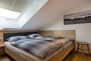 Ein Schlafzimmer mit Oberlicht, einem Bett mit Karodecke, einem Holz-Kopfteil, einem Nachttisch, einem Holzschemel und einem schwarz-weißen Landschaftsbild an der Wand.