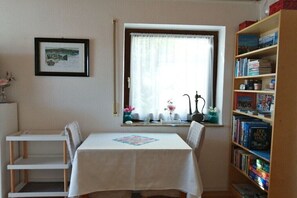 Una habitación con una mesa, sillas, una estantería, un ventanal con cortinas y un cuadro en la pared.