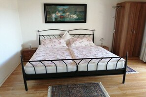 Una cama con estructura metálica, una colcha estampada con motivos florales, una mesita de noche con lámpara, un armario de madera y un cuadro en la pared.
