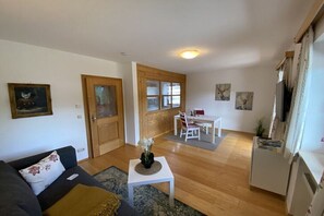 Ein Wohnzimmer mit einer Couch, einem kleinen Tisch mit einer Vase, einem Essbereich mit Tisch und Stühlen sowie einer Küchenzeile mit Kühlschrank.