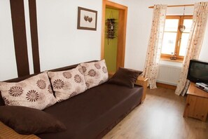 Ein Wohnzimmer mit braunem Sofa, einem Fernseher auf einem Holzschrank, einem Fenster mit Vorhängen und einer Tür mit hängender Dekoration.
