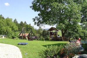 Ein Spielplatz mit Schaukeln und einer Rutsche, umgeben von Bäumen und einer Rasenfläche.