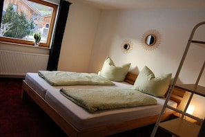 A bed with a wooden frame, two pillows, a bedside table with a lamp, and a window with a view of a building and trees. - Holiday home with 3 bedrooms (Oberstdorf \/ Tiefenbach)
